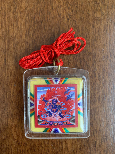 A clear square shaped amulet of the deity, Nam Jon. A red thread is attached to the amulet by a metal ringlet so it could be worn. The image of Nam Jon inside the amulet and decorated with thread details on the back. The amulet has smooth edges.