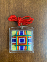 Load image into Gallery viewer, The back of the amulet showing the thread detailing - it is incased in the plastic casing so the front and back of the amulet are smooth. There are five colors (green, orange, red, white, and blue) woven into a square design.