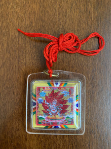 A clear square shaped amulet of the deity, Nam Jon. A red thread is attached to the amulet by a metal ringlet so it could be worn. The image of Nam Jon inside the amulet and decorated with thread details on the back. The amulet has smooth edges.