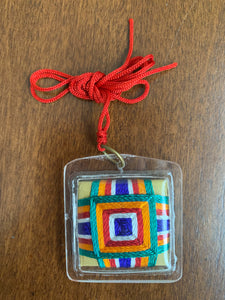 The back of the amulet showing the thread detailing - it is incased in the plastic casing so the front and back of the amulet are smooth. There are five colors (green, orange, red, white, and blue) woven into a square design.